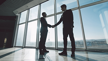 Two businessmen in suits shaking hands in front of large floor-to-ceiling windows in a bright, modern office.