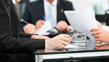 A close-up of professionals in suits sitting around a glass table, reviewing and signing legal documents during a meeting.